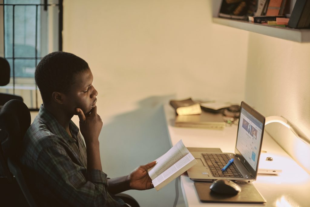 Male student reading a book, sitting at a desk with a laptop