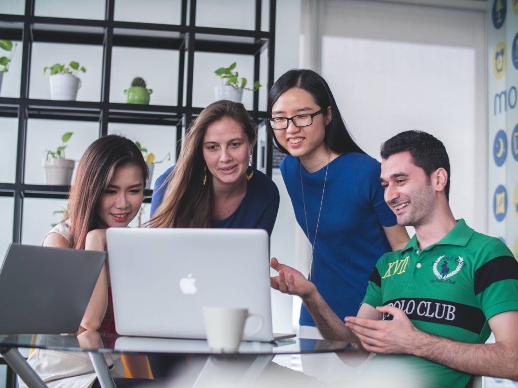 Three female students and one male study stand around a laptop working on a task together